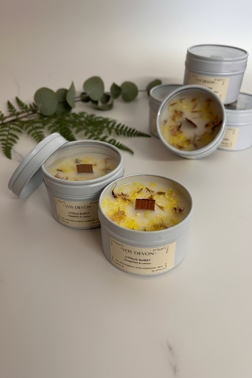 Several round metal candle tins with cream labels are arranged on a plain beige surface. Two tins are open, showing a creamy off-white wax with yellow dried botanical pieces and a small brown bark accent, while others are closed.