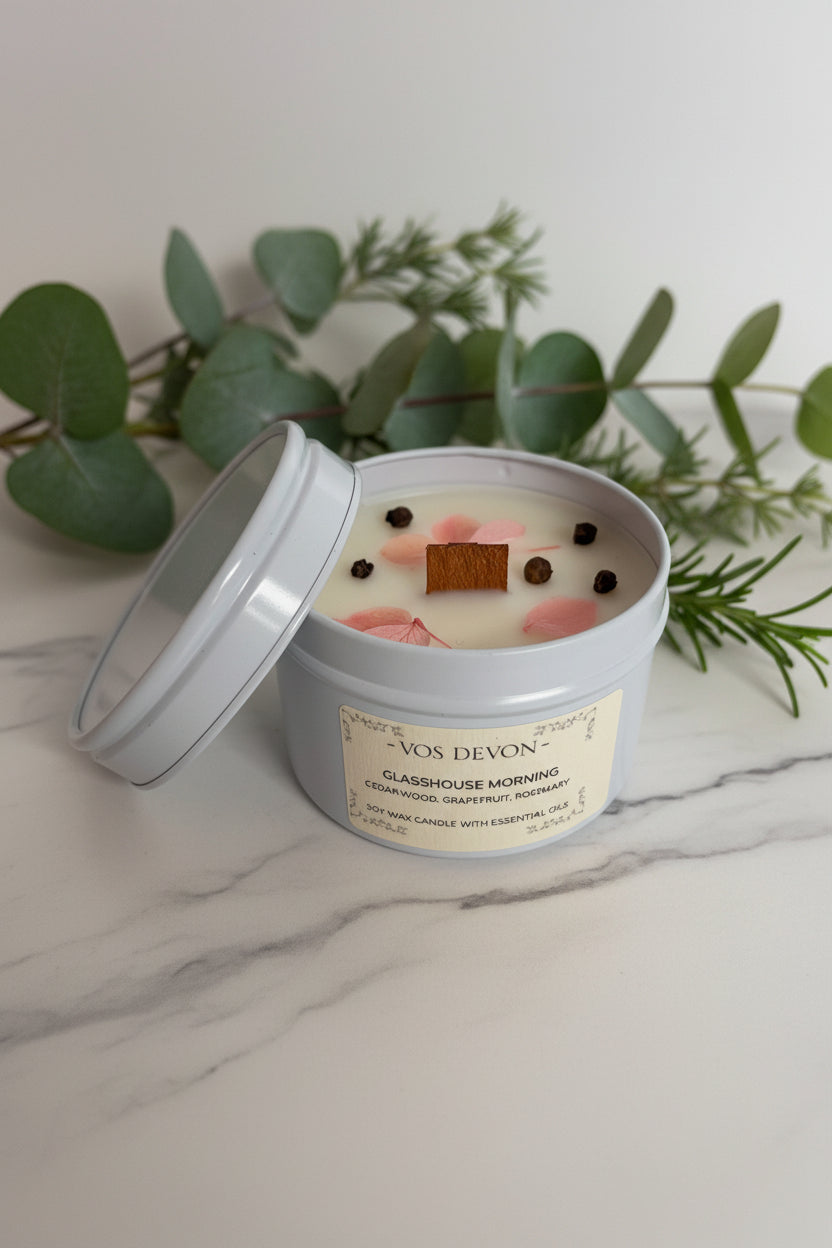 A VOS DEVON 'Glasgow Morning' scented candle in a light grey tin with its lid propped open, featuring pink flower petals and dark peppercorns on a cream-colored wax surface.