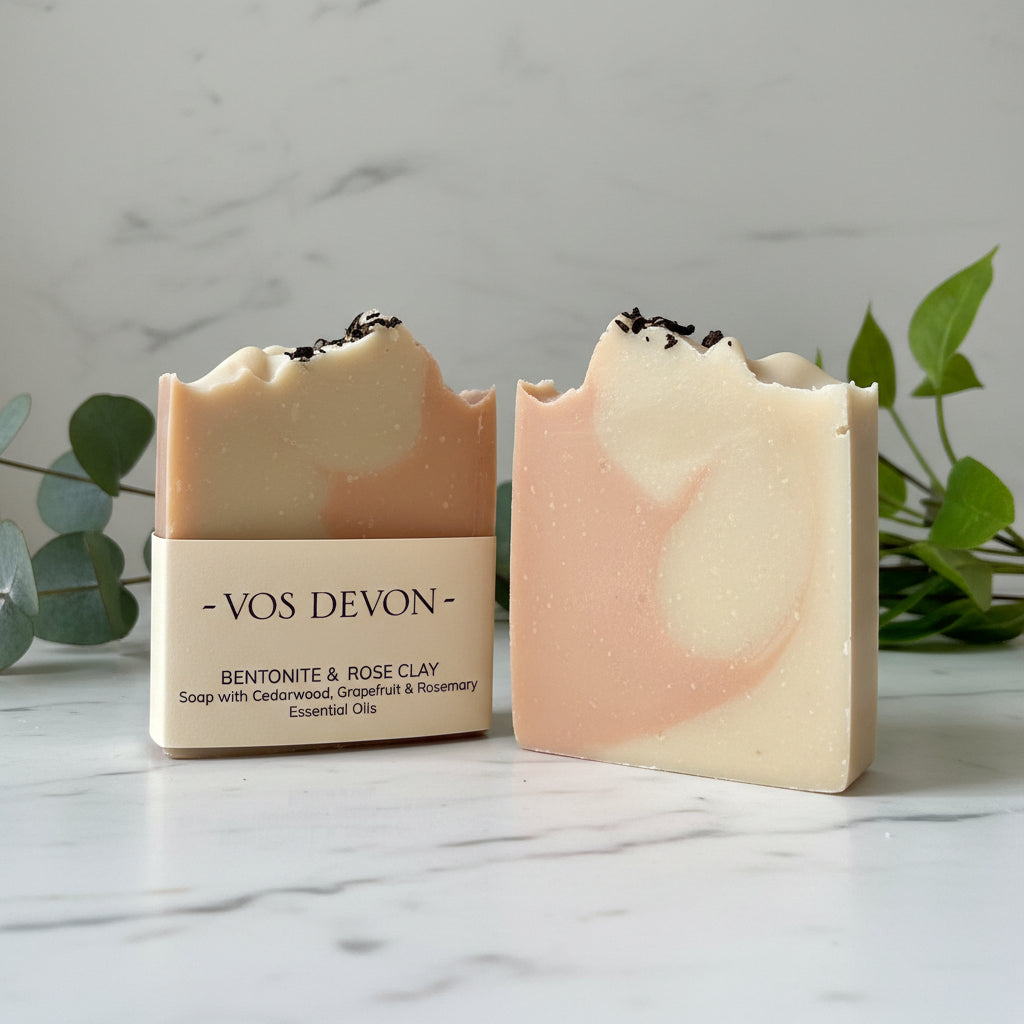 Two handmade rectangular bars of soap with rough, broken tops, featuring a marbled pattern of peach-pink and cream. One bar has a paper label reading 'VOS DEVON - BENTONITE & ROSE CLAY'.