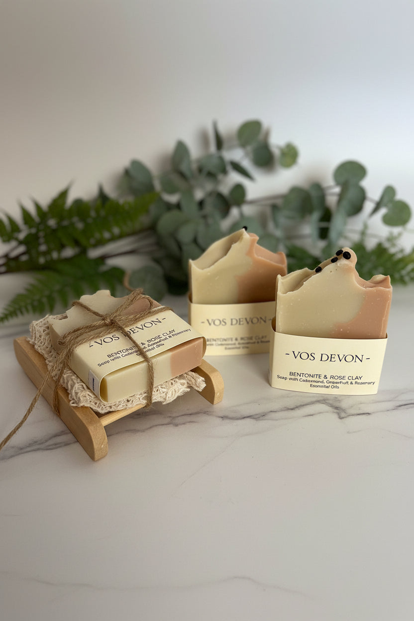 Four handmade soap bars with animal-shaped tops are displayed on a neutral background, including two wrapped in paper bands labelled 'VOS DEVON' and one wrapped in a lace sleeve tied with twine on a small wooden stand.