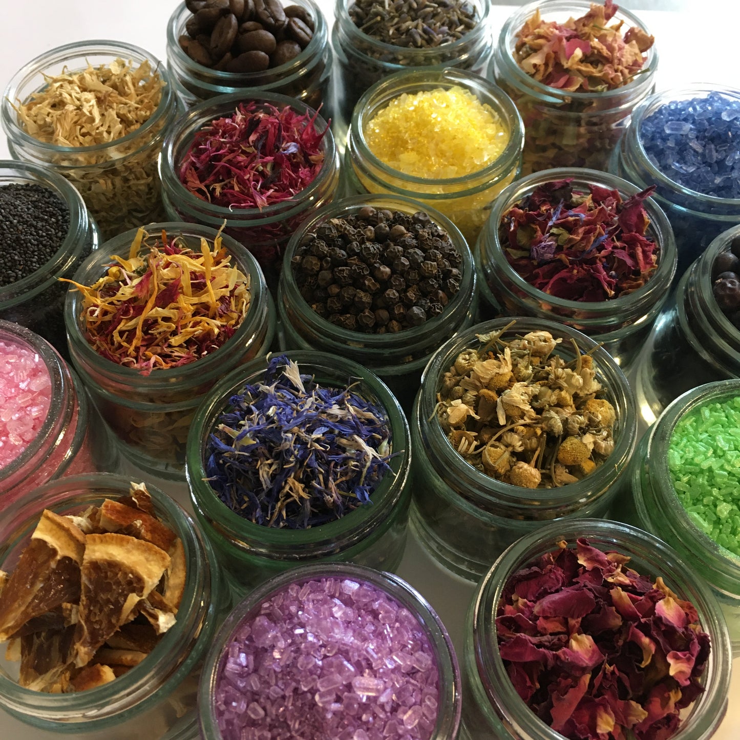An overhead close-up of many small clear glass jars filled with colourful dried botanicals and crystals, including pink, purple, yellow, blue, brown, black, and red tones.