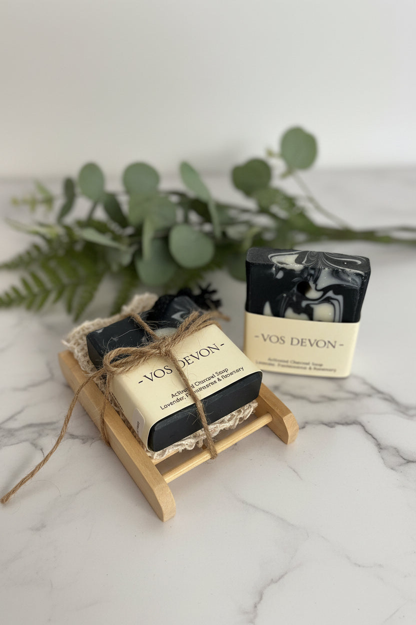 Two bars of VOS DEVON soap displayed on a small wooden stand against a plain light background. One bar is wrapped in cream paper with jute twine, and the other stands upright in its matching cream wrapper.
