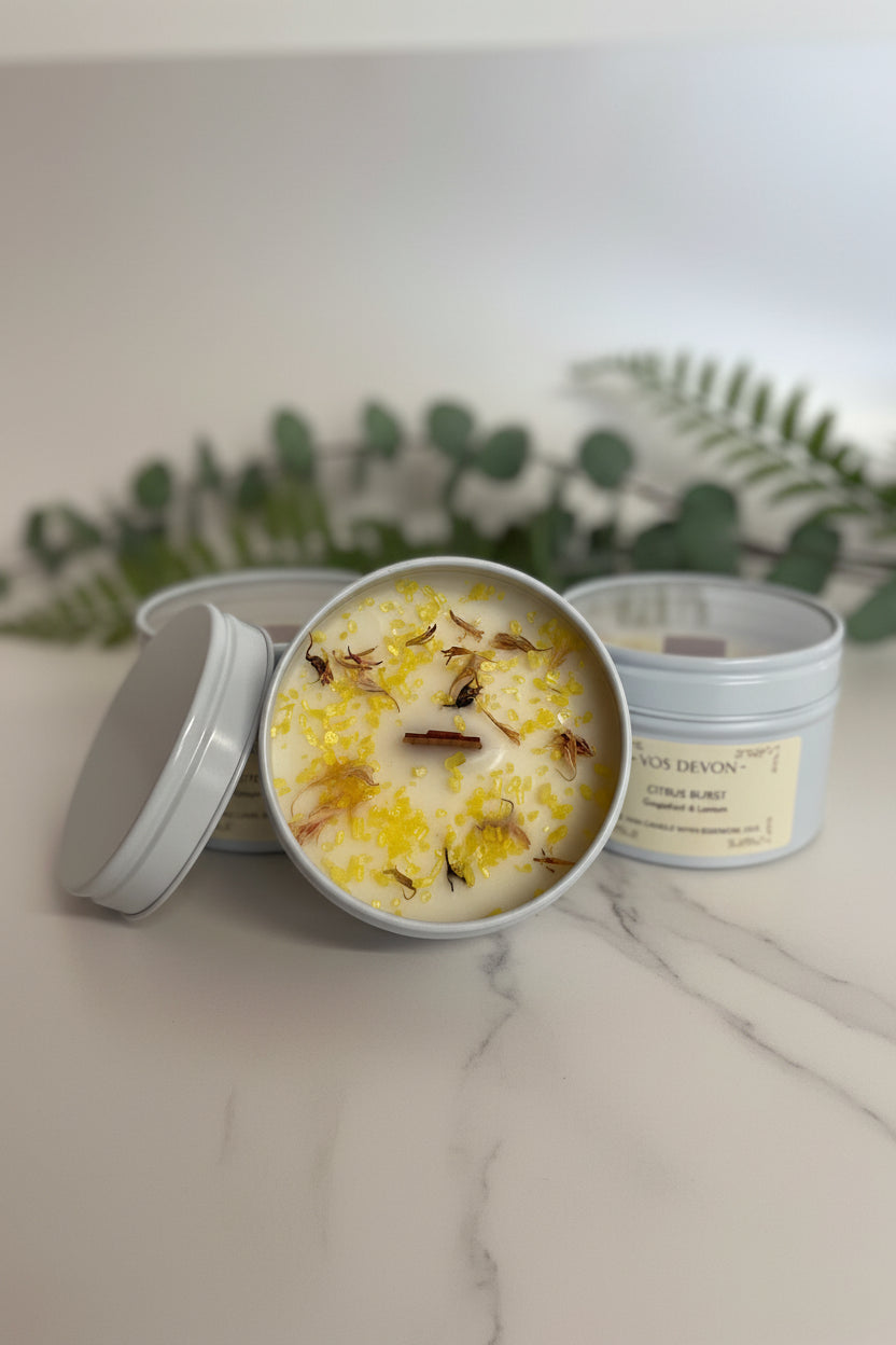 Three round metal candles in light grey tins on a warm beige surface. The center tin is open and shows creamy off-white wax topped with yellow petals and small dried botanical pieces.