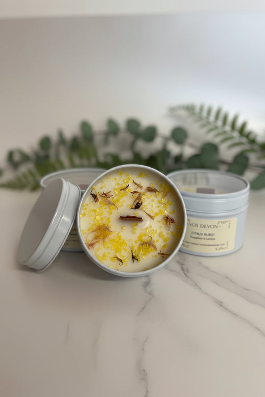 Three round metal candles in light grey tins on a warm beige surface. The center tin is open and shows creamy off-white wax topped with yellow petals and small dried botanical pieces.