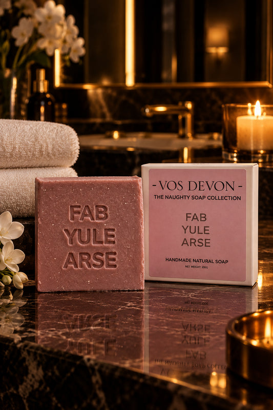A pink handmade soap bar embossed with 'FAB YULE ARSE' sits on a marble surface next to its pink and white packaging box from VOS DEVON.