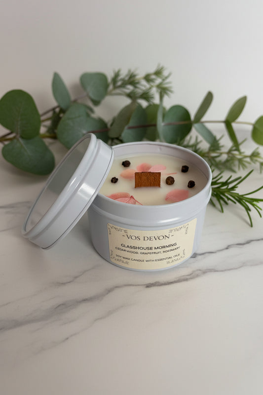A VOS DEVON 'Glasgow Morning' scented candle in a light grey tin with its lid propped open, featuring pink flower petals and dark peppercorns on a cream-colored wax surface.