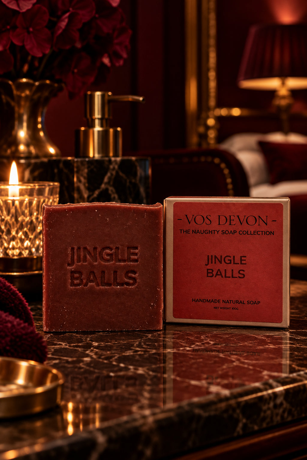 A bar of brown handmade soap embossed with 'JINGLE BALLS' stands next to its red and white paper packaging box on a marble table. The box is branded 'VOS DEVON - THE NAUGHTY SOAP COLLECTION'.
