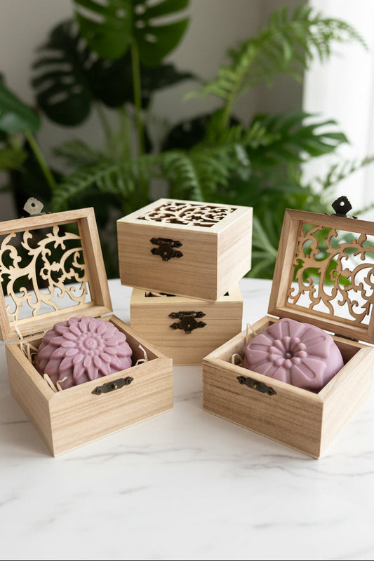 Three light natural wood gift boxes with ornate laser-cut wooden lids and small metal clasps, arranged on a white surface. Two open boxes contain round dusty mauve-coloured soaps nested in wood wool.