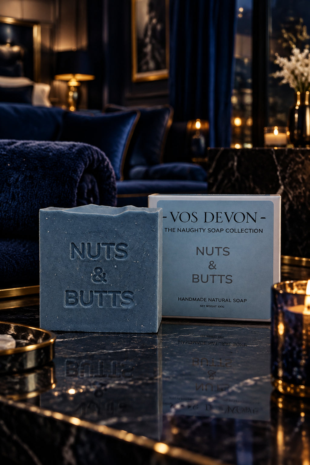 A rectangular bar of Nuts & Butts handmade natural soap in a muted slate blue-grey colour, standing next to its light blue and white branded packaging box on a marble table top.