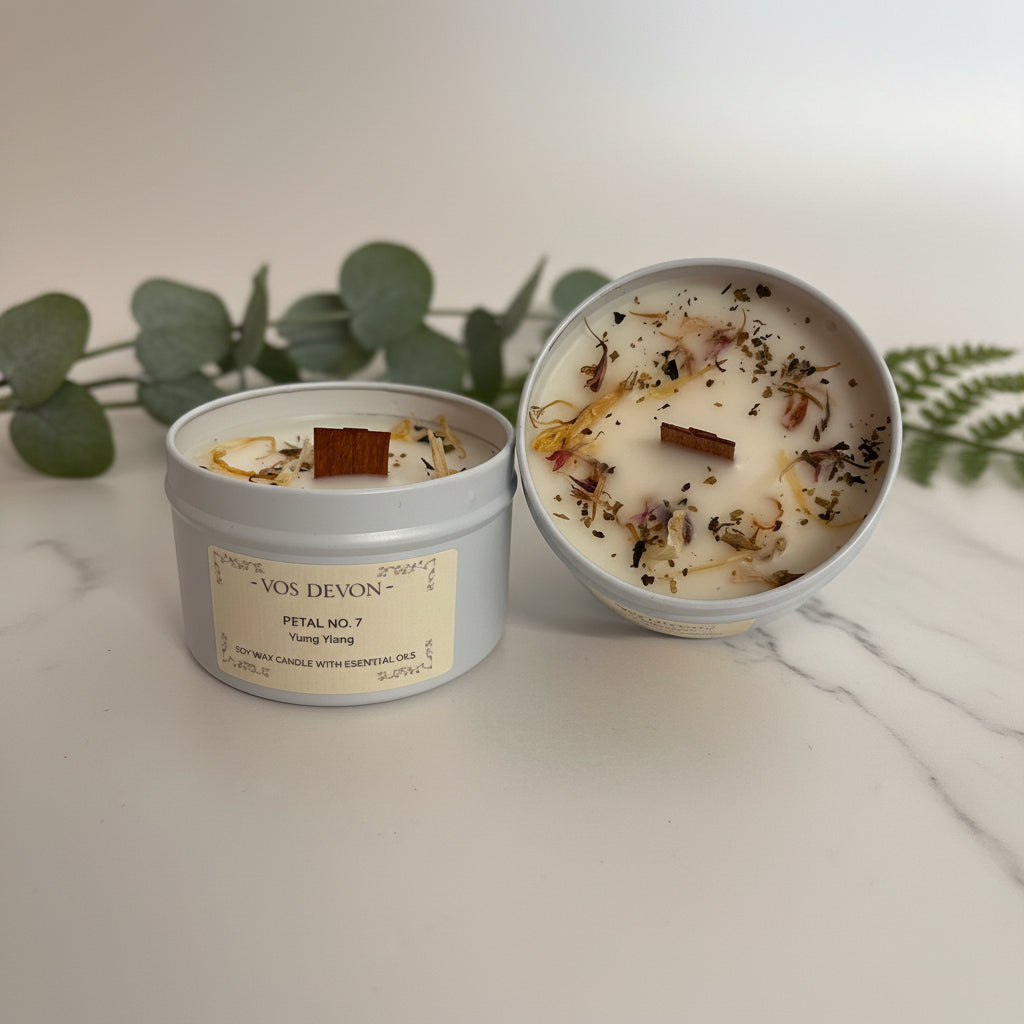 Two round candles in light grey metal tins on a plain light background. One tin is closed with a cream label reading “VOS DEVON” and “PETAL NO.