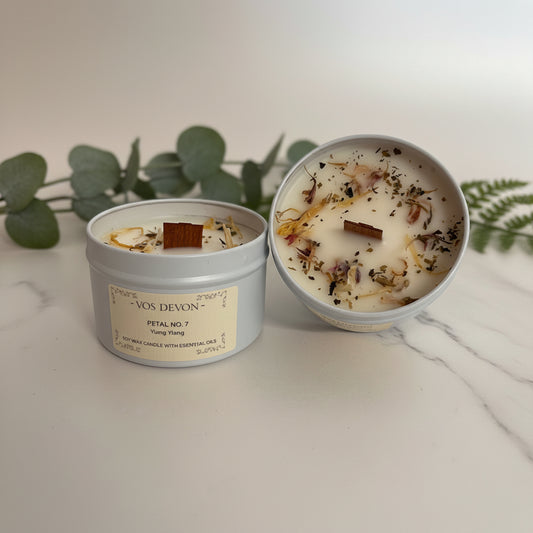 Two round candles in light grey metal tins on a plain light background. One tin is closed with a cream label reading “VOS DEVON” and “PETAL NO.
