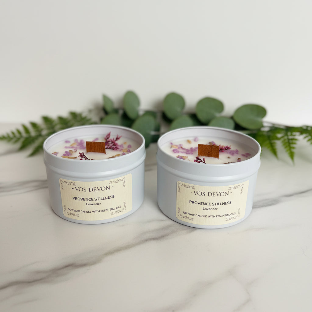 A white soy candle in a light grey container with a cream label, featuring dried lavender buds and a small cinnamon stick on top, photographed on a plain off-white background.
