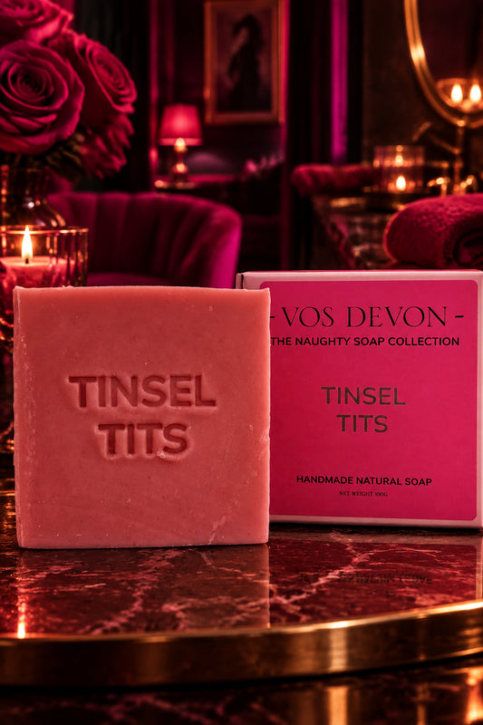 A rectangular bar of dusty pink soap embossed with 'TINSEL TITS' sits in front of its matching pink packaging box on a marble surface. The box reads 'VOS DEVON - THE NAUGHTY SOAP COLLECTION' and 'TINSEL TITS'.