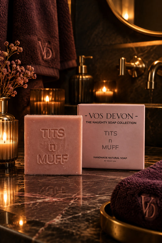 A pink handmade soap bar embossed with 'TITS n MUFF' stands in front of its matching pink packaging box on a marblen surface. The box reads 'VOS DEVON - THE NAUGHTY SOAP COLLECTION' and 'TITS n MUFF'.