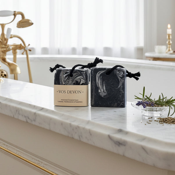 Two rectangular bar soaps with black hanging cords, one wrapped in a beige paper label reading 'VOS DEVON - Activated Charcoal Soap', shown against a light grey background.