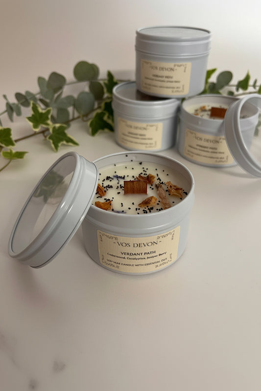 Four silver metal tins of candles with cream labels are arranged on a light beige surface. One tin in the foreground is open, showing white wax topped with small black seeds and brown bark pieces, while the other tins stand closed behind it.